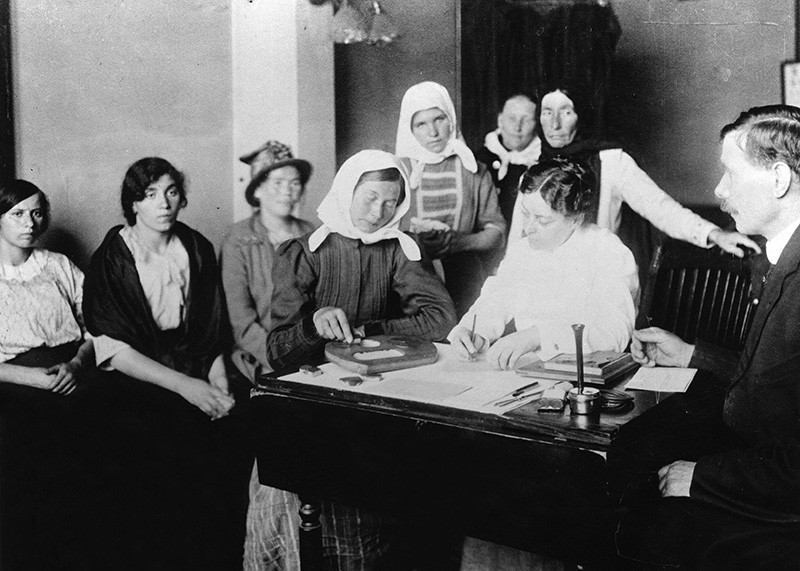Black and white photo of a woman doing a puzzle at a desk, with other people waiting around her.