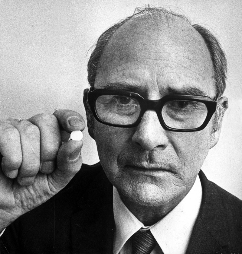 Black and white picture of John Cade in a jacket, tie and glasses, holding a white pill.