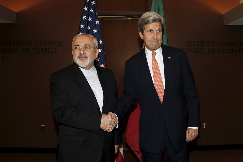 John Kerry shakes hands with Mohammad Javad Zarif. The US and Iranian flags are in the background.