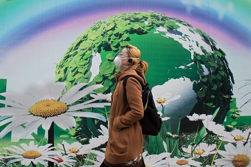 A woman wearing a protective pollution mask walks past a billboard depicting a green globe and flowers in Beijing
