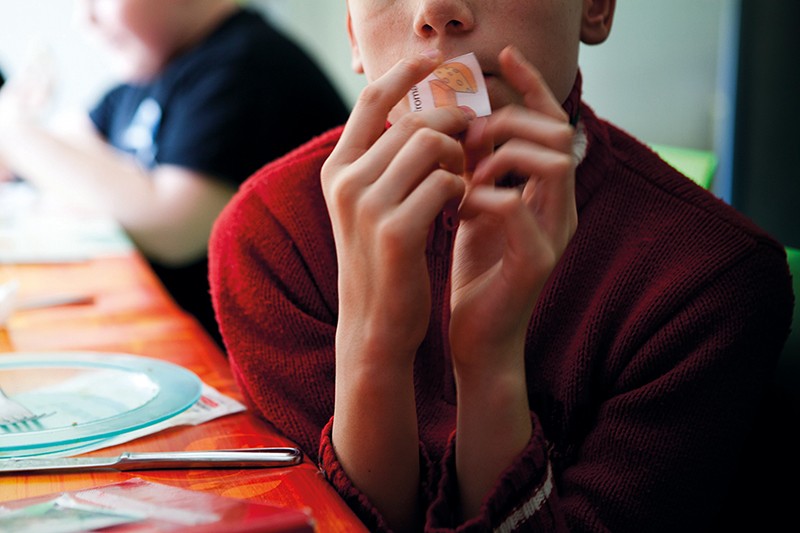 A child at a school dinner table holds up a small piece of paper with a symbol on it