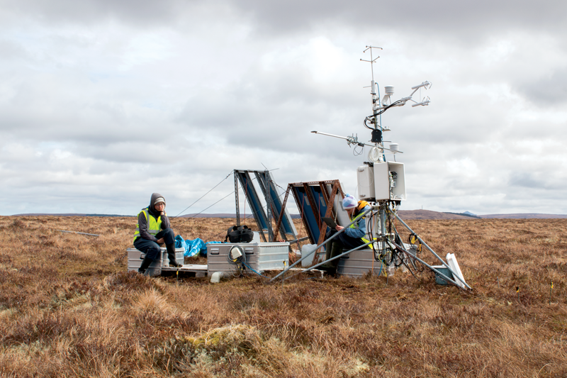 Two researchers sit surrounded by monitoring equipment that make up a Flux tower on an empty blanket bog