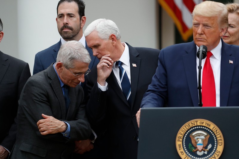 Mike Pence whispers to Anthony Fauci while Donald Trump stands at a lectern during a press conference