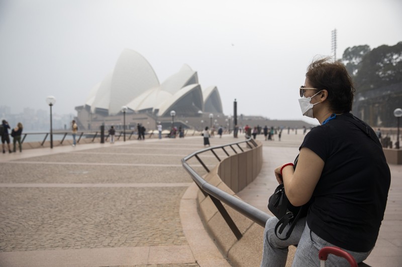 A woman wearing a face mask sits near the Sydney Opera House shrouded in haze