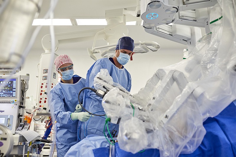 Two medics in full scrubs stand by a da Vinci robot