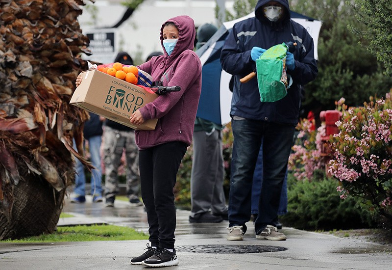 A recipient carries a box of food as others wait in line at a Food Bank distribution the sidewalk.