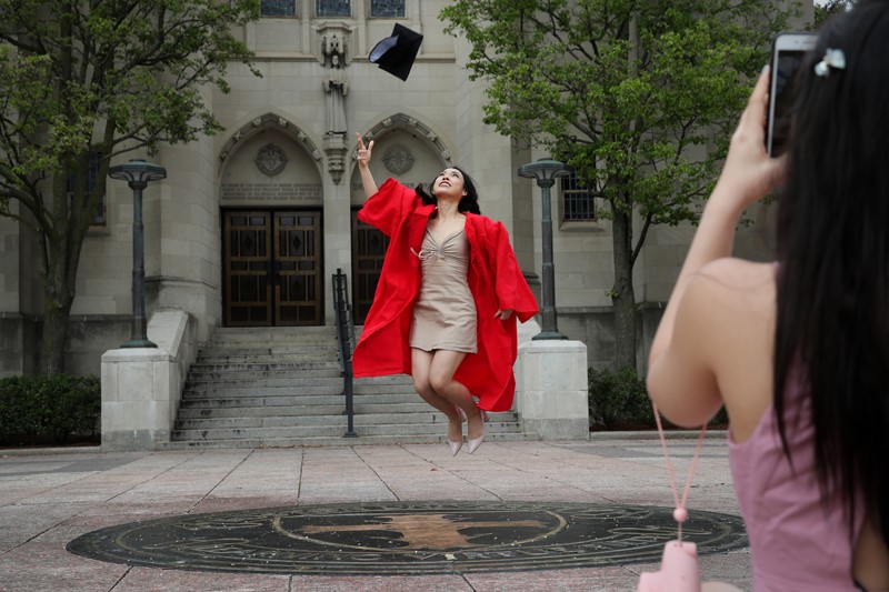 A graduate wearing a red gown throws a mortarboard into the air and jumps while someone takes her photograph