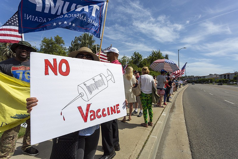 A protester on a roadside holds a sign depicting a syringe labelled '666', with the words 'No Vaccine' in red.
