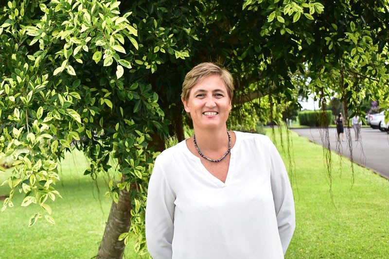 A woman in a white top, standing under a tree, smiles at the camera.