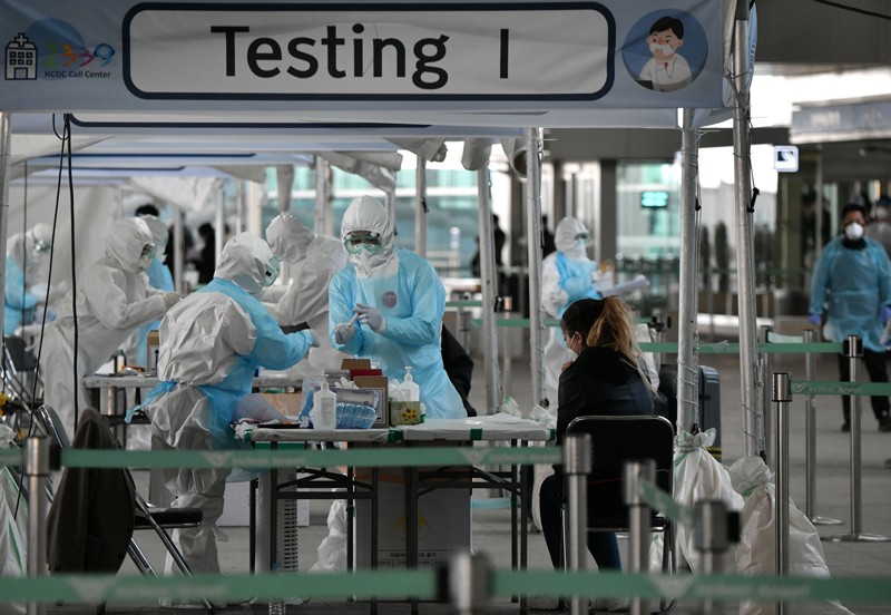 Medical staff wearing protective clothing take test samples from a traveller in a covered area marked 'Testing'