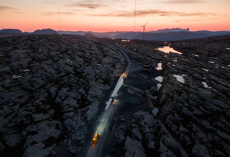 A large wind turbine blade is transported by road through a rocky landscape at sunset with a wind turbine in the distance