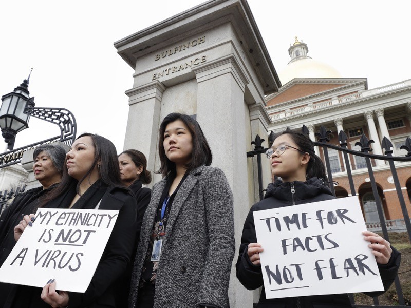 Several women stand outside a building. Two hold signs that say 'My ethnicity is not a virus' and 'Time for facts not fear'.