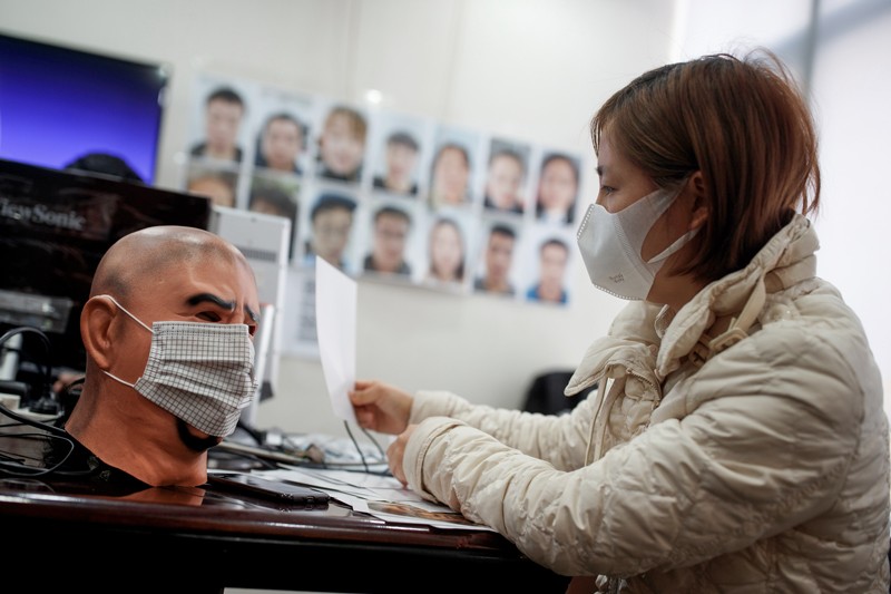 A woman wearing a face mask sits at a desk next to a mannequin head wearing a face mask with portraits hung on the wall behind