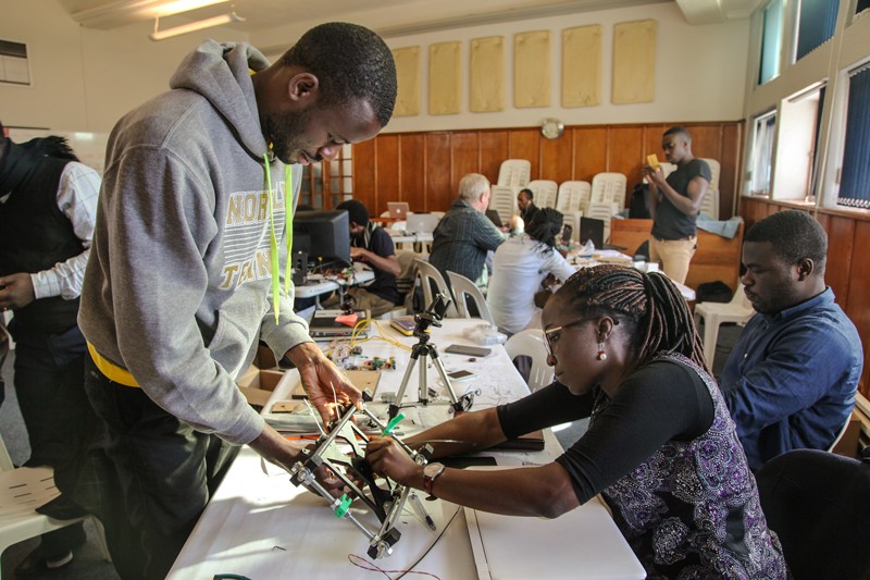 Two people work at a table on a metal and plastic frame while other people work at tables around them