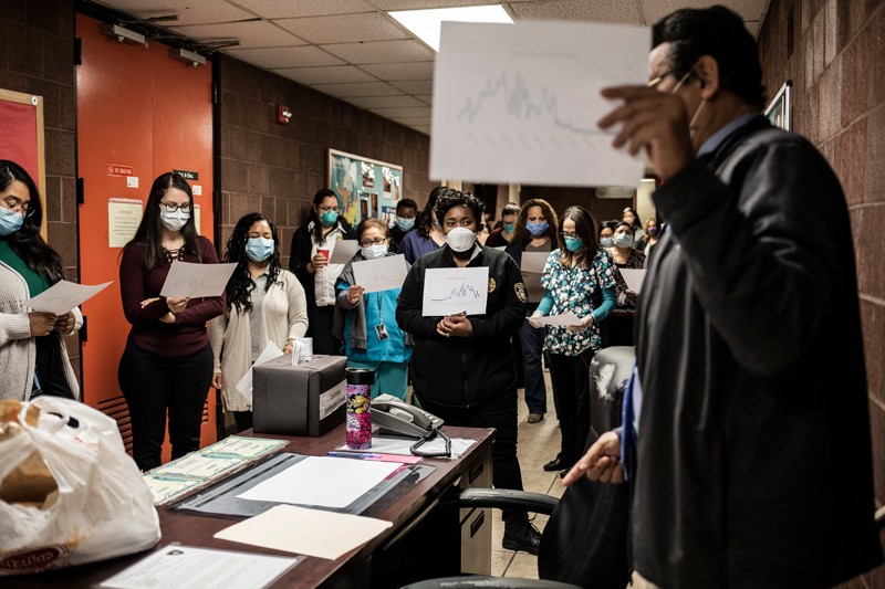 In an office, a man holds up a piece a paper showing a chart to a group of people in masks working as contact tracers