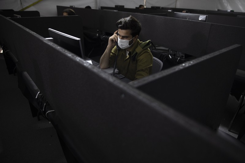 A man in army fatigues and a face mask sitting in a booth at a computer talking on the phone