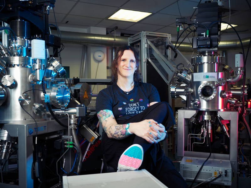 Clara Barker sitting in a lab surrounded large metallic lab equipment and wearing shoes with blue, pink and white striped soles