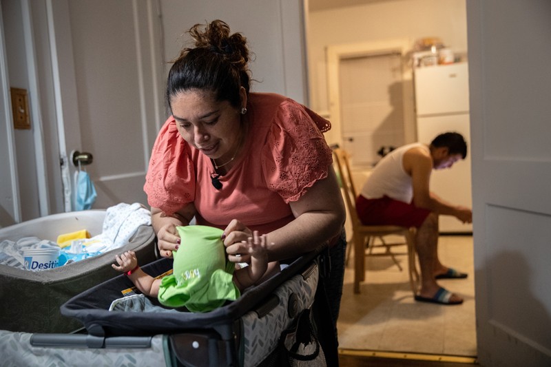 A woman puts clothes on a new born baby while a man sits on a chair in the room behind