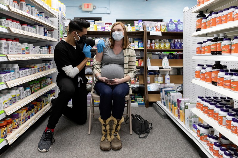 A heavily pregnant woman wearing a face mask receives a vaccine whilst sitting in the aisle of a pharmacy