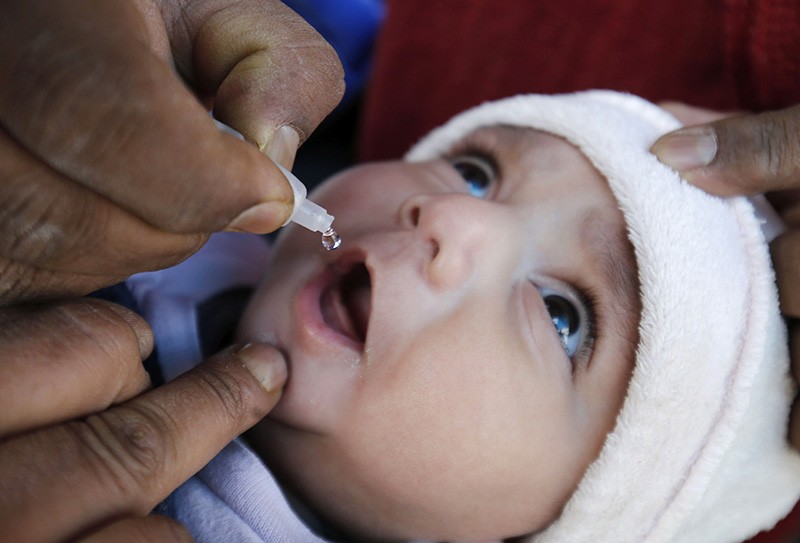 Close up of a child's face. Two hands hold their mouth open and a third squeezes a drop of vaccine from a bottle.