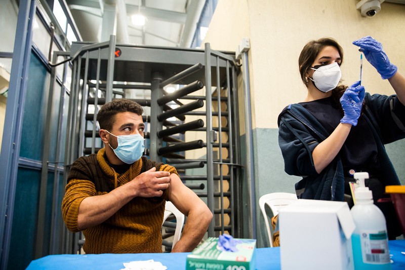 A man in a mask rolls up his sleeve as a health worker prepares a syringe.