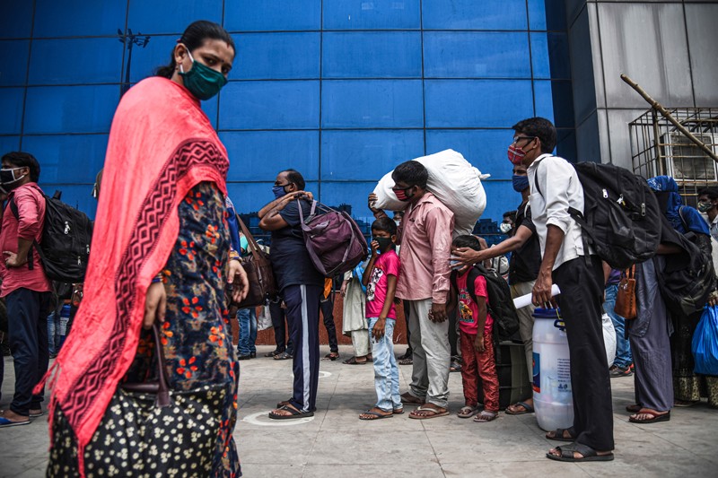 Migrant farmers wearing face masks and carrying luggage queue at a railway station.