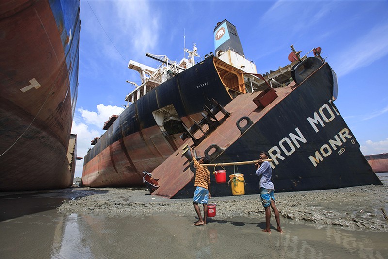 Two people carry buckets next to large ships grounded on a beach.