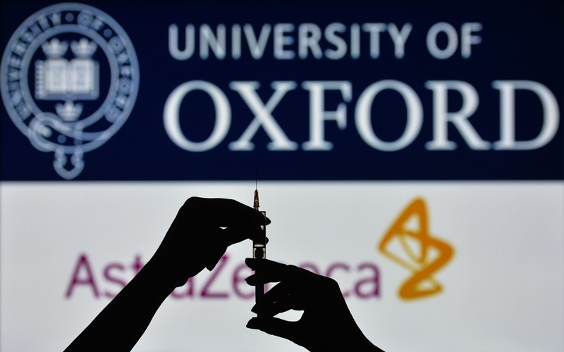 A silhouette of hands holding a medical syringe in front of the Oxford University and AstraZeneca logos