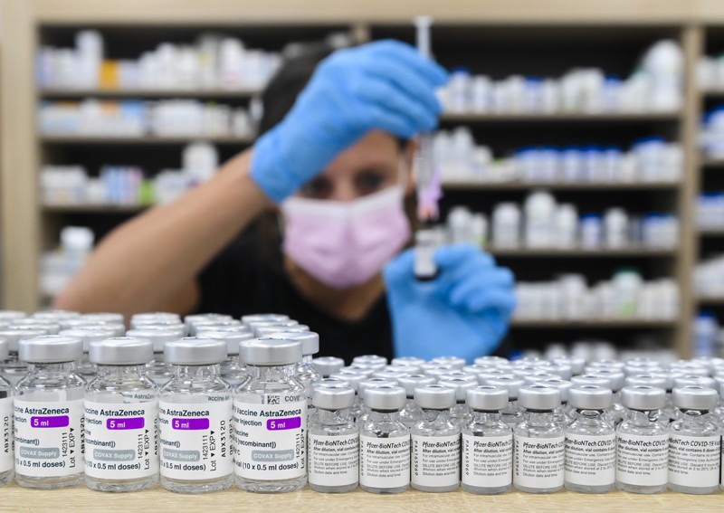 A pharmacy worker prepares a vaccine while sitting behind a shelf stacked with AstraZeneca and Pfizer COVID-19 vaccine vials