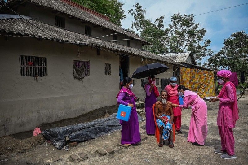 Five female health workers wearing pink and purple attend to an elderly man receiving a COVID-19 vaccine in rural India.