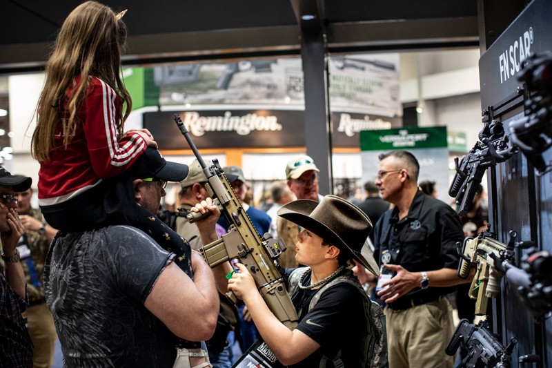 A young girls sits on a man's shoulders as a boy handles an assault rifle at a gun convention in Texas