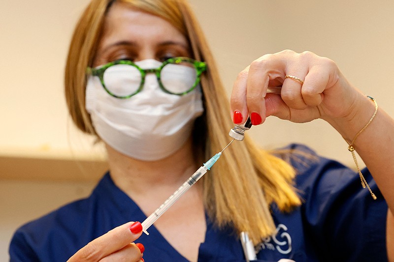 A woman wearing a mask and glasses uses a syringe to extract the vaccine from a vial.