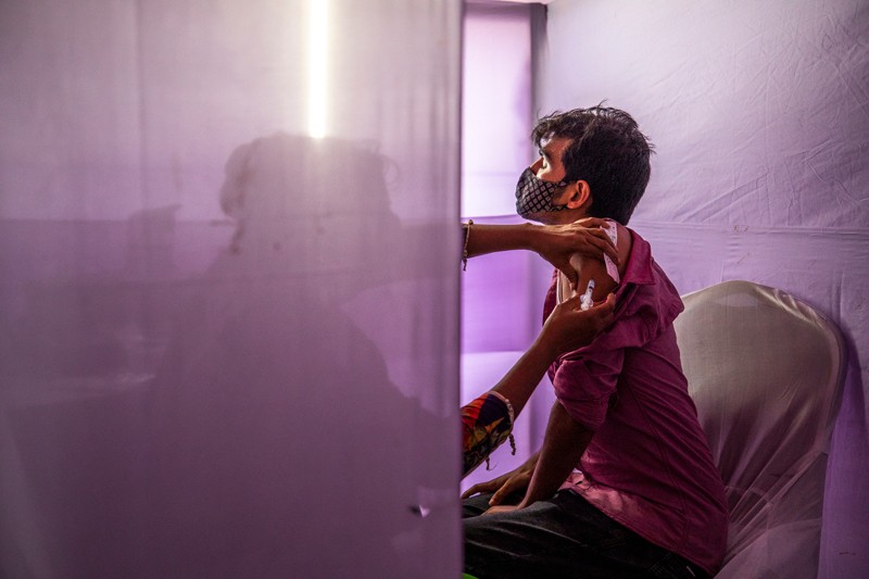 A health worker silhouetted behind a purple sheet administers a COVID-19 vaccine to a man in Bangladesh