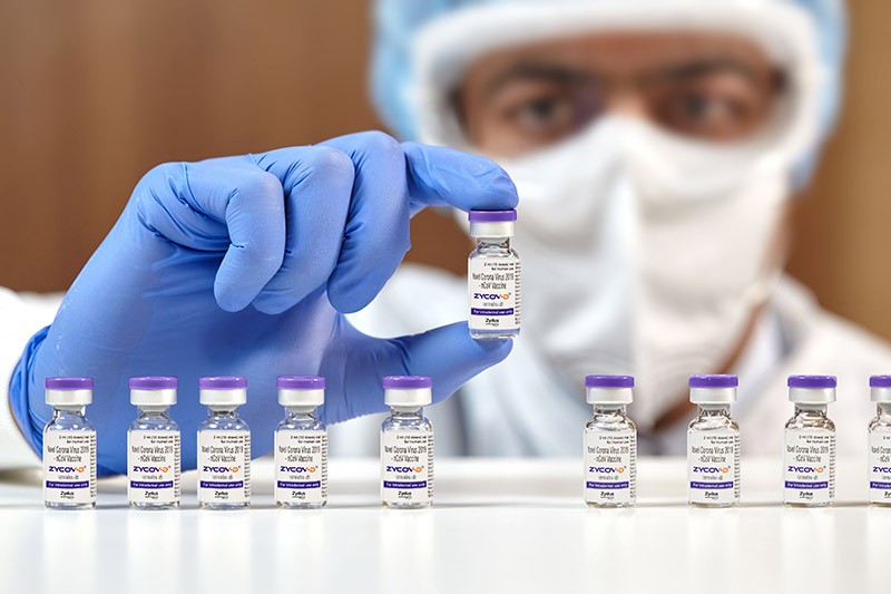 A person in protective clothing holds a single vial above a display of many vials of ZyCoV-D.