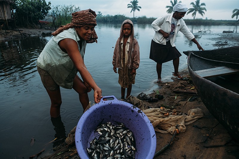 Three people standing by water, one is displaying their catch of fish, in the coastal town of Maroantsetra in Madagascar