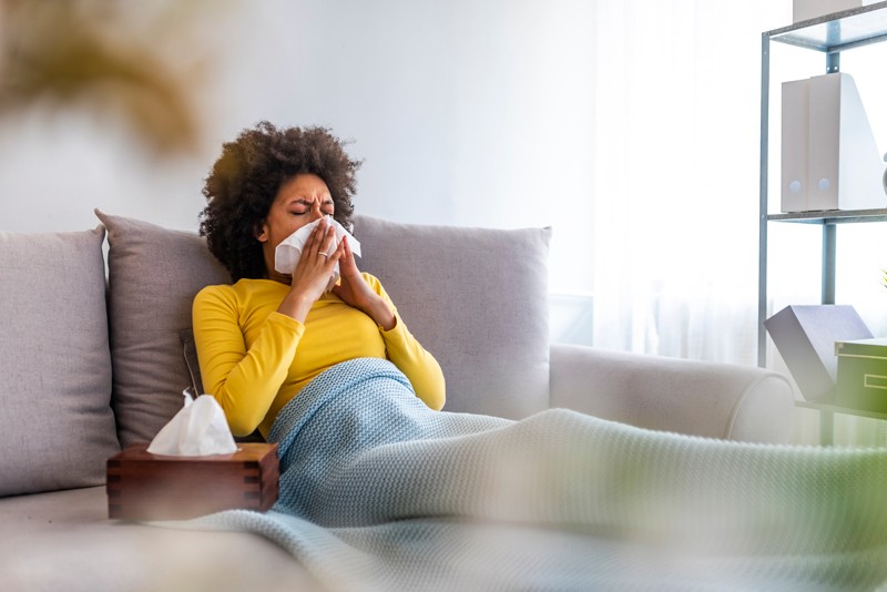 A sick young woman sneezing into a tissue at home