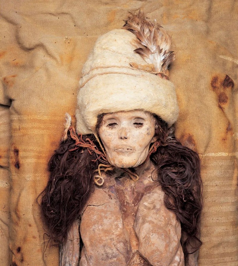 The remains of a naturally mummified woman with hair, wearing a white hat with a feather in it.