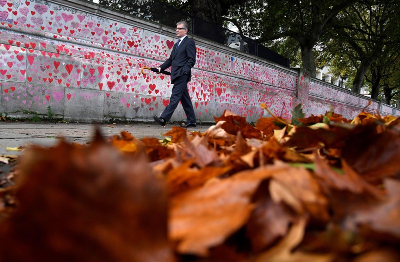 A man walks through fallen leaves beside a wall covered in hand-drawn hearts and messages commemorating COVID victims in London