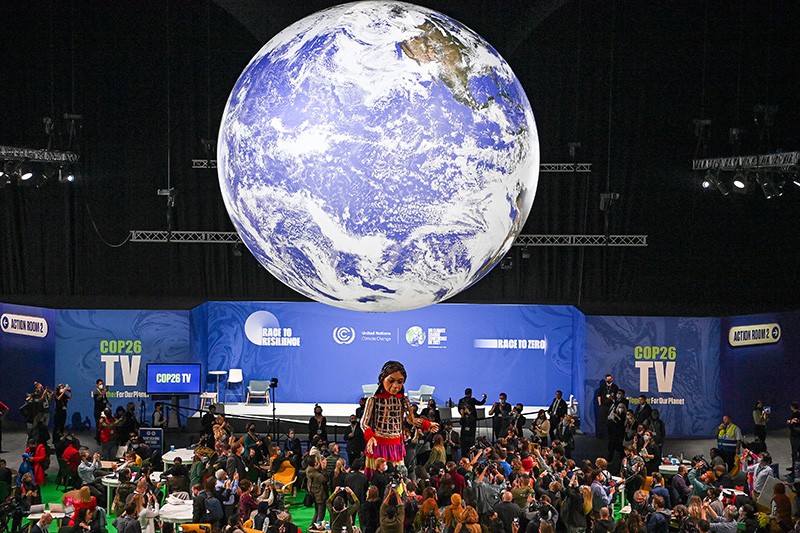 A giant Earth and a puppet depicting a Syrian refugee girl is seen at COP26 along with attendees and signage.