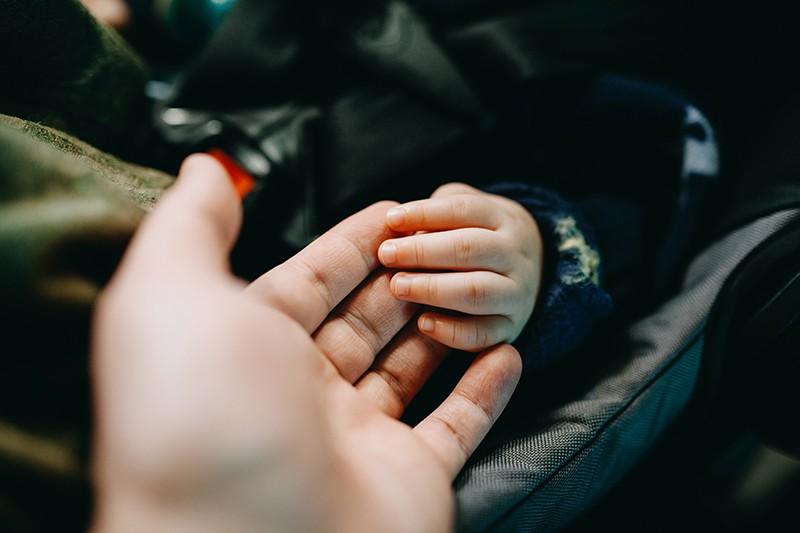 Close up of a father holding a baby's hand gently, while the baby is in a car seat.