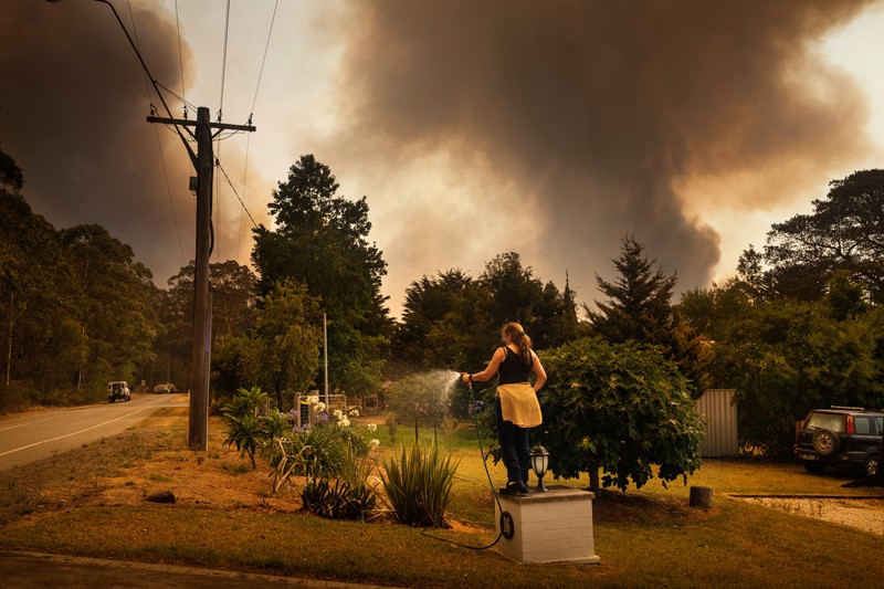 A woman waters her garden with a hose as plumes of black smoke rise from an approaching bush fire in Australia