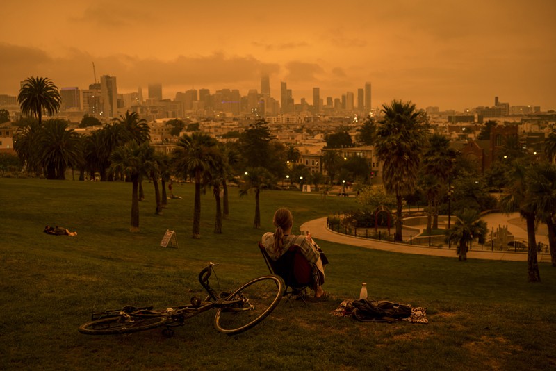 A person sits in a park overlooking San Francisco with a sky yellow from smoke and fog