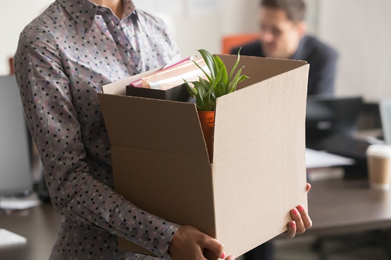 A person carrying a cardboard box with office supplies and a plant in an office.
