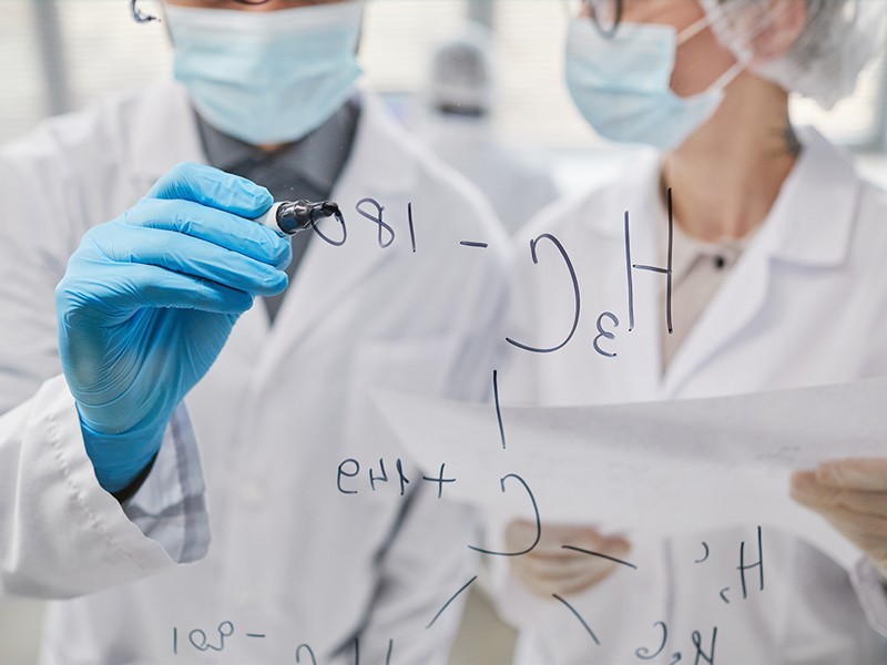 Close-up of two scientists writing chemical formulas on glass board and discussing them in team.