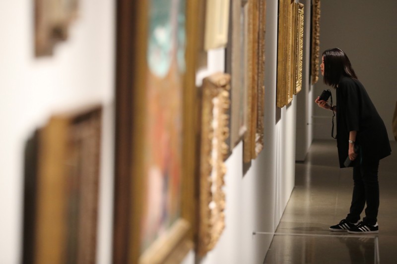 A gallery visitor looks closely at a painting hanging on the wall next to many other framed paintings