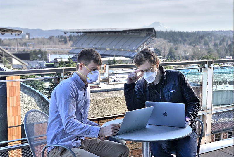 Wearing facemasks, Alex DeGrave and Joseph Janizek sit a table on a balcony looking at their laptops