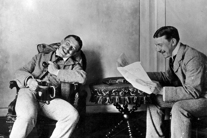 Black and white photo of two men laughing while one reads from a newspaper circa 1920