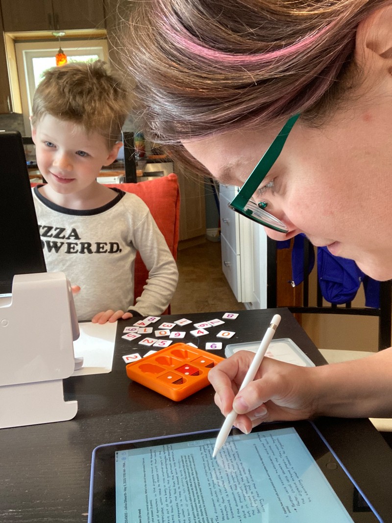 Cynthia Harley works on a tablet computer while her child looks at a computer and plays with numbered tiles