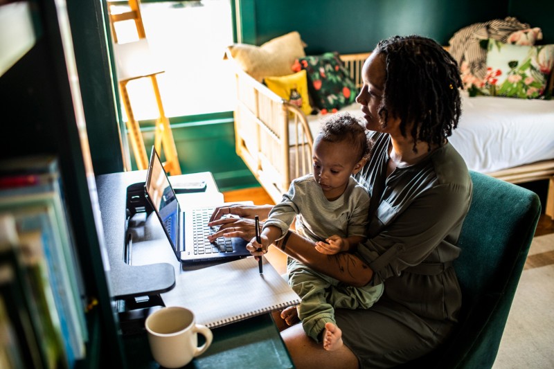 Woman working from home on a laptop while holding a toddler who is drawing in a notebook