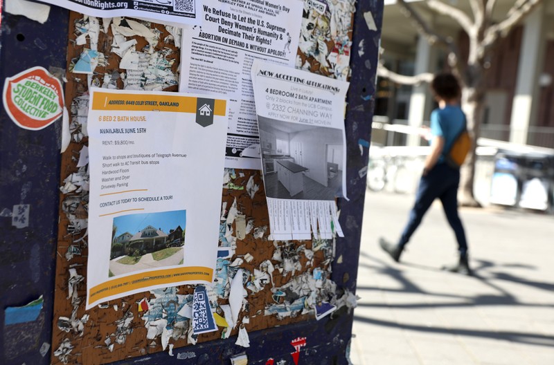 A young man walks past a notice board with signs for home rentals on a university campus